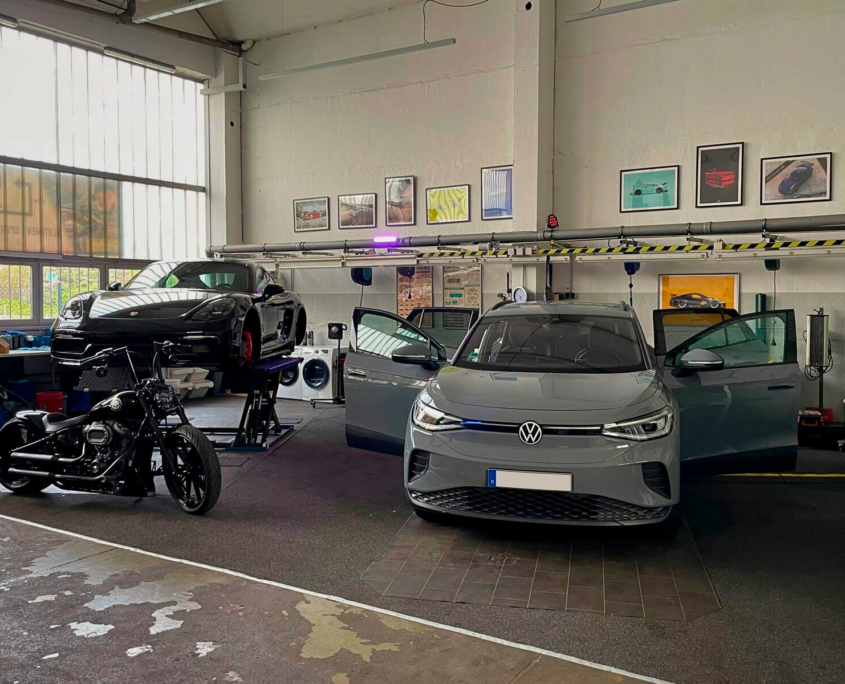Dresden-Gutschein-Distrex-03 Helle, moderne Werkstatthalle mit grauem VW im Vordergrund, einem schwarzen Porsche auf einer Hebebühne und einem schwarzen Motorrad.
