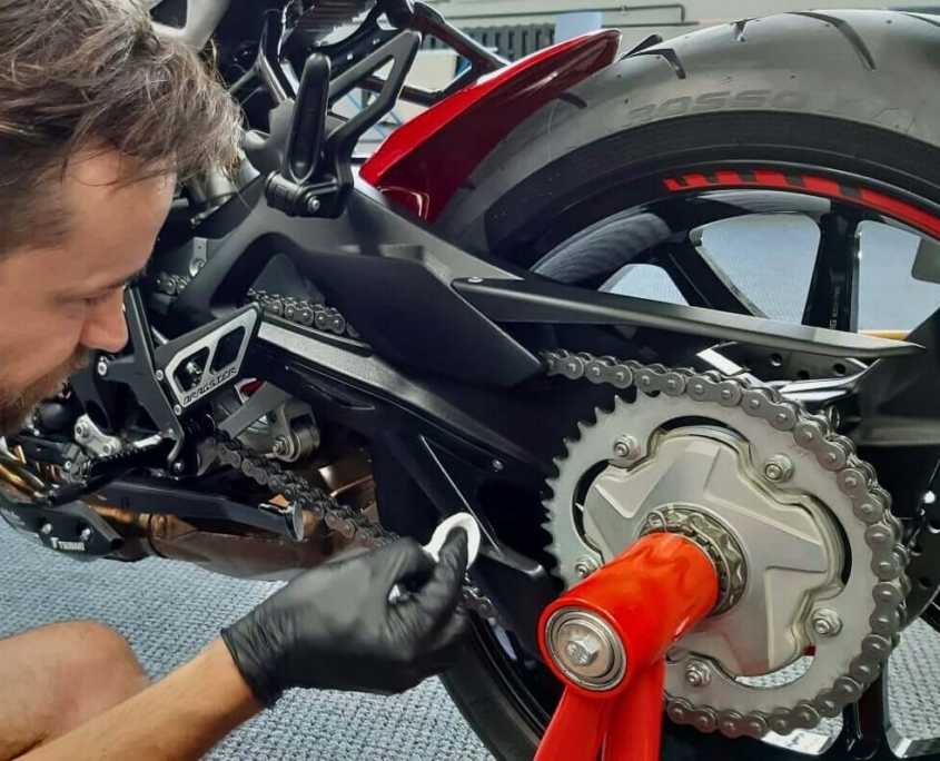 Dresden-Gutschein-Distrex-07 Nahaufnahme eines Mechanikers mit schwarzen Handschuhen, der die Antriebskette und das Kettenrad eines Motorrads präzise reinigt oder fettet.