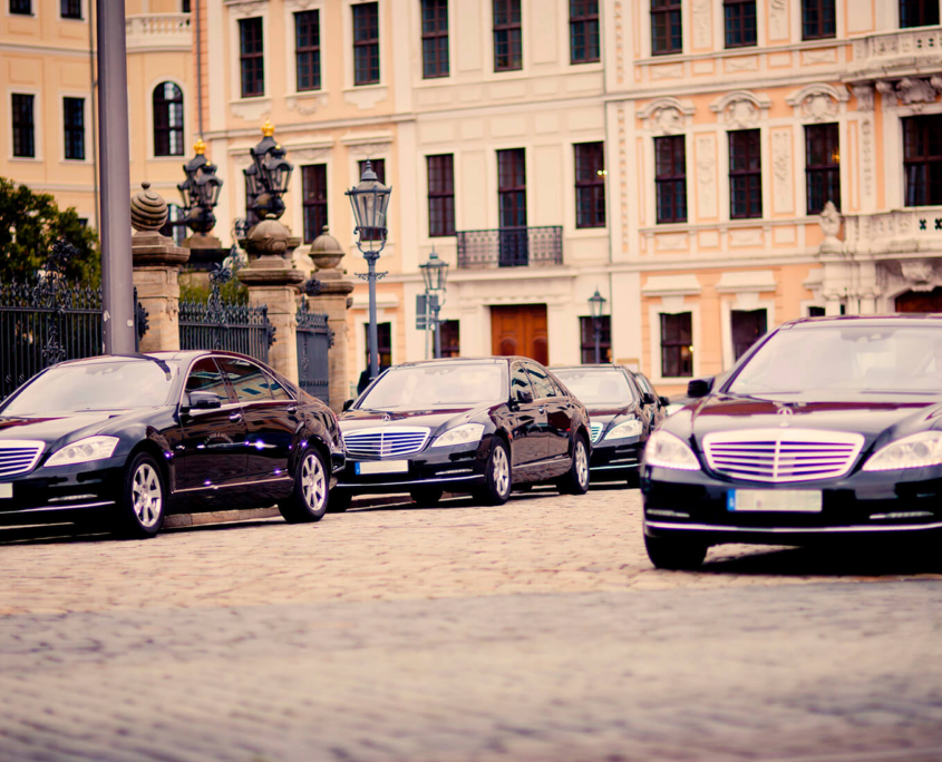 Mehrere identische, schwarze Mercedes-Benz Fahrzeuge sind in einer Reihe auf einem historischen Platz hintereinander aufgestellt.