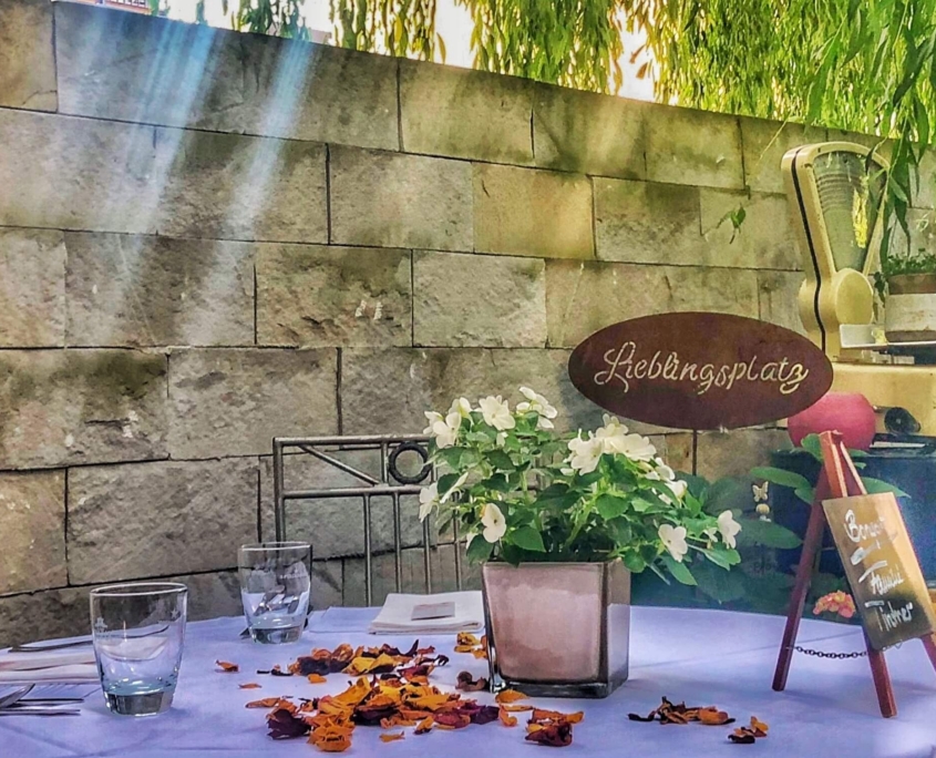 Außenansicht eines liebevoll gedeckten Tisches vor einer rustikalen Sandsteinmauer mit einem Schild „Lieblingsplatz“ und Blumendekoration im Sonnenlicht.