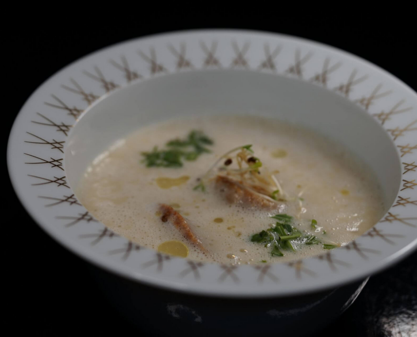 Nahaufnahme einer fein angerichteten, schaumigen cremeweißen Suppe mit Kräutern und Einlage in einem edlen Porzellanteller.