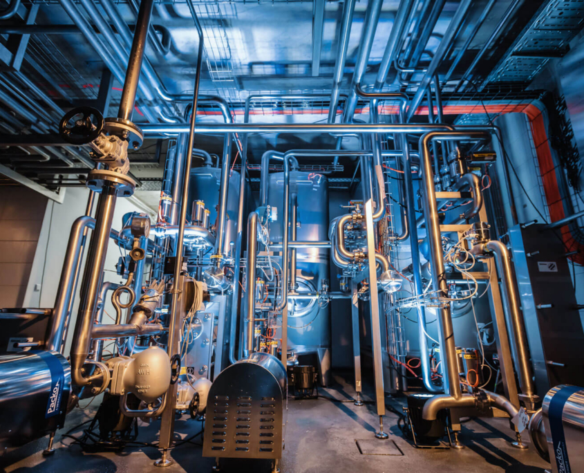 Dresdner Whisky Manufaktur 03 Hochmoderne Destillationsanlage der Dresdner Whisky Manufaktur mit komplexen Edelstahlsäulen und Rohrleitungssystemen in atmosphärischer blauer Beleuchtung.